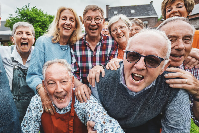 A lively group of over-65 friends gathered at a countryside house, laughing and shouting together as they take a joyful group selfie.